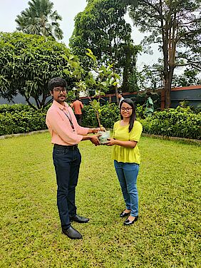 A man and a woman in a large garden holding a potted sapling 