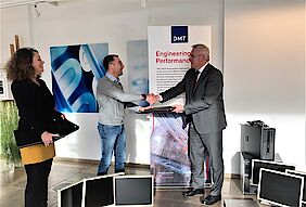 2 men, surrounded by computers and monitors, shake hands as one hands a laptop to the other.  